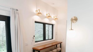 Una sala de estar moderna con piso de madera, un sofá de cuero negro, una mesa de comedor de madera con cuatro sillas y un ventanal con vista a árboles.