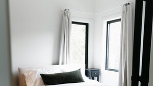 Un dormitorio con cama, mesita de noche, dos ventanas con cortinas y una alfombra estampada.