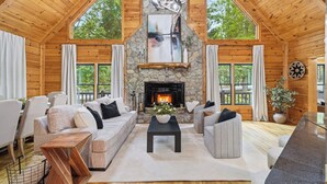 A log cabin interior with a stone fireplace, wooden ceiling, and large windows.