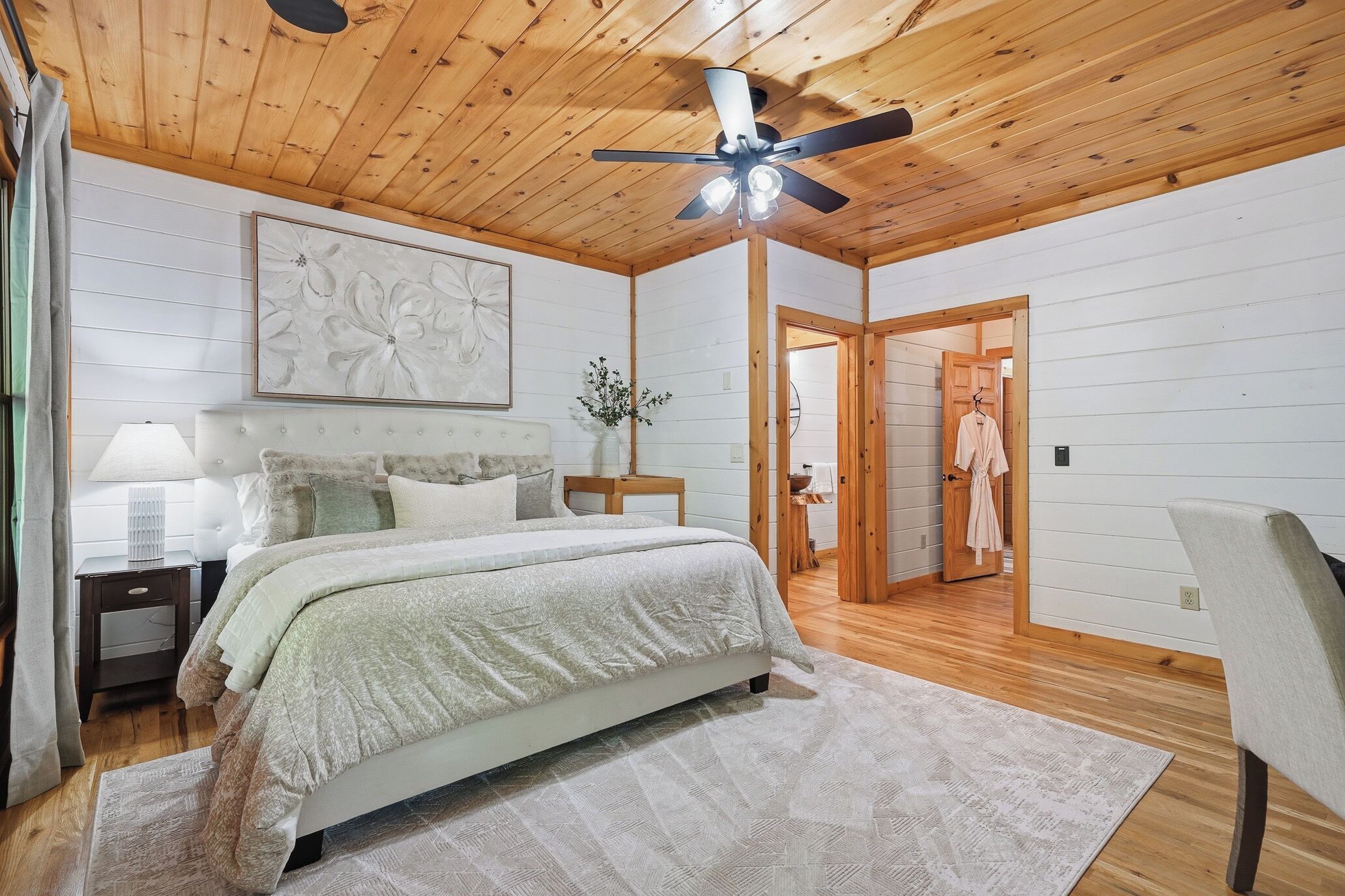 Une chambre à coucher comprenant un lit, un ventilateur de plafond, une table de chevet, une lampe, un tableau et une porte donnant sur une autre pièce.