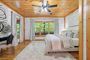 A bedroom with a bed, a desk, a chair, a ceiling fan, and a window with curtains.
