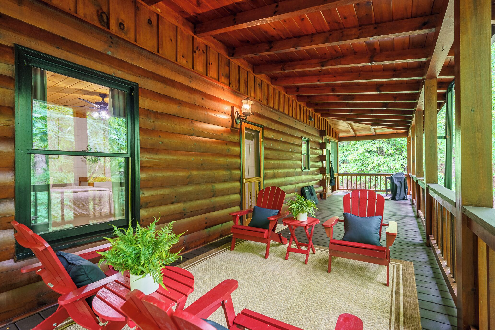A log cabin with a porch, red Adirondack chairs, a small table with a plant, and a view of the forest.