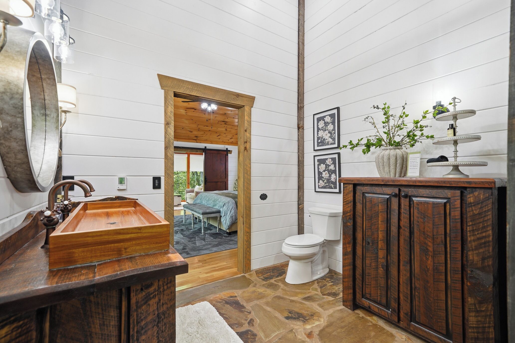 A bathroom with a wooden vanity, a large mirror, a sink, and a toilet. There is a door leading to a bedroom.