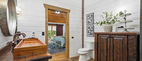 A bathroom with a wooden vanity, a large mirror, a sink, and a toilet. There is a door leading to a bedroom.