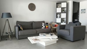 A modern living room with a grey sofa, a coffee table, a lamp, and a bookshelf. - Friendly Beach House (Sandy Ground)