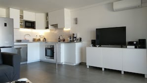 A modern kitchen with white cabinetry, a built-in oven, a microwave, and a coffee maker. There is a flat-screen TV on a white cabinet and a speaker on the floor. - Friendly Beach House (Sandy Ground)