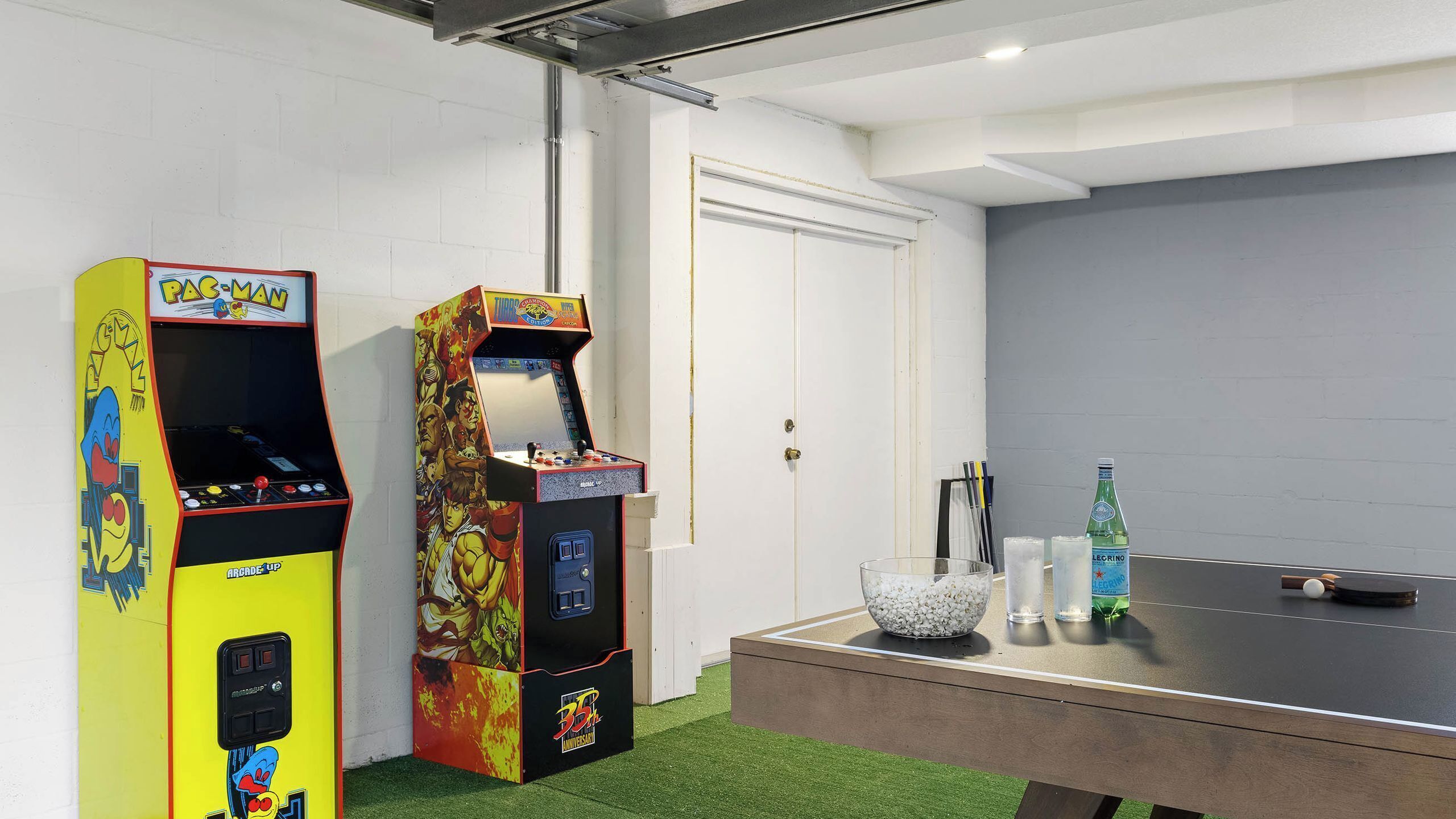 Deux bornes d’arcade, une table de ping-pong et une porte dans une pièce aux murs blancs et au tapis vert.