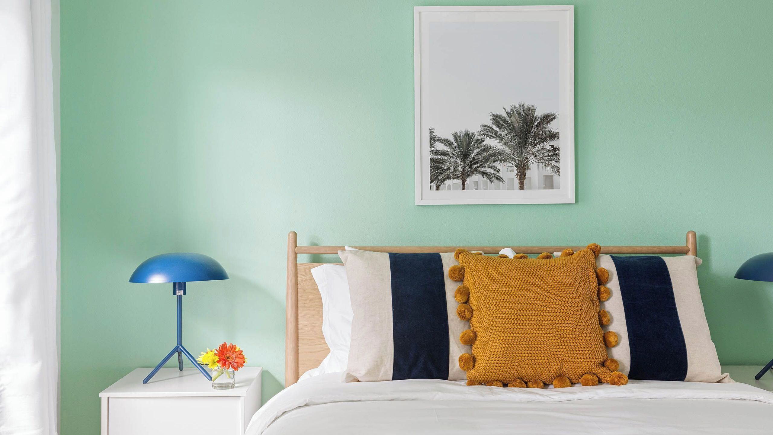 A bedroom with a bed, a nightstand with a lamp, a framed picture on the wall, and a vase with flowers.