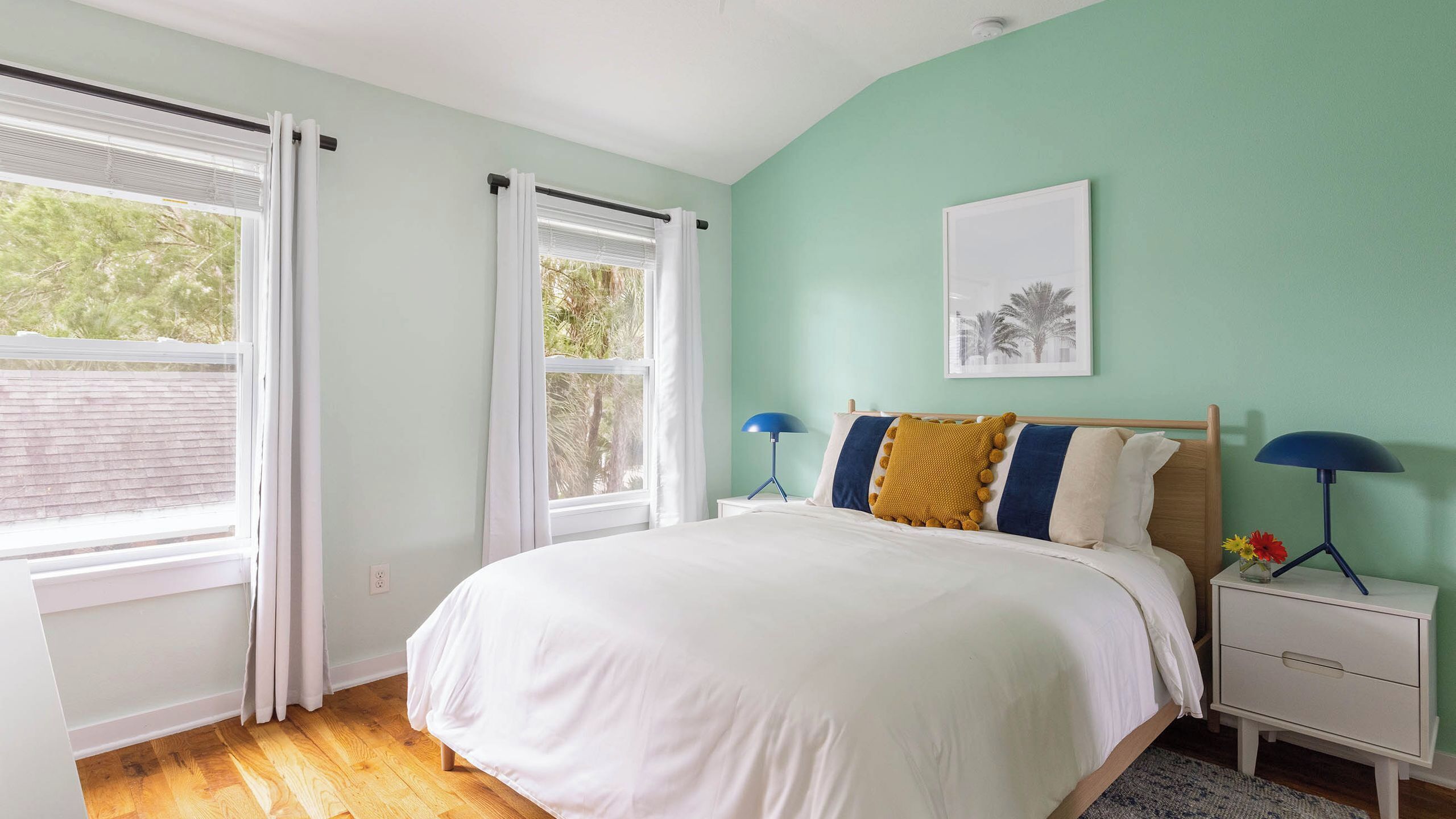A bedroom with a bed, two bedside tables, a window with curtains, a wall-mounted picture, and a wooden floor.