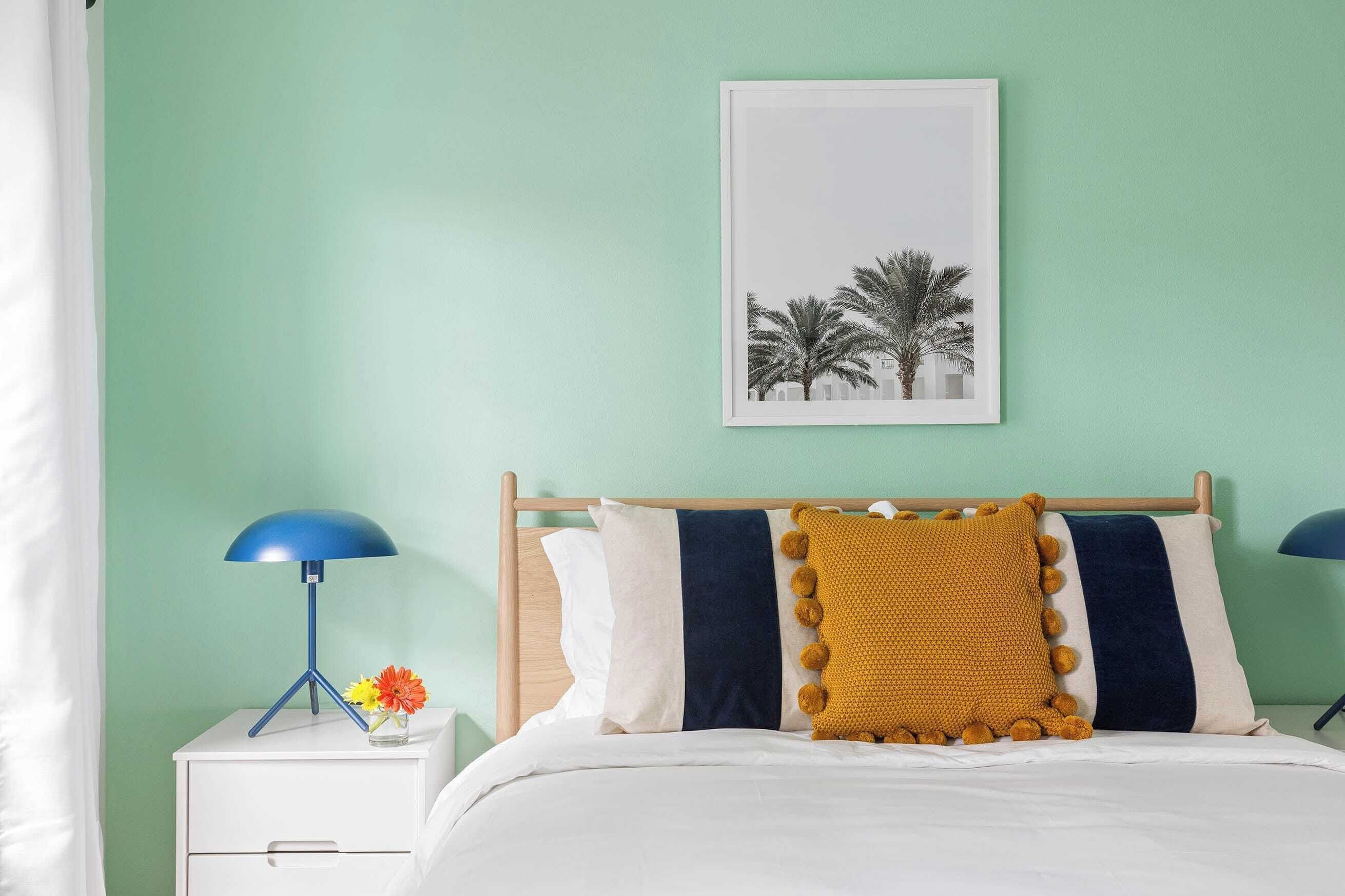 A bedroom with a bed, a nightstand with a lamp, a framed picture on the wall, and a window with curtains.