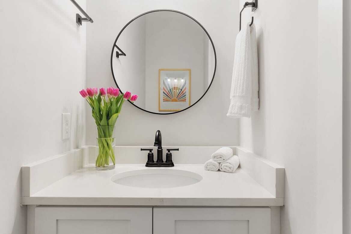 A bathroom with a white sink, a round mirror, a framed picture, and a vase with pink flowers.