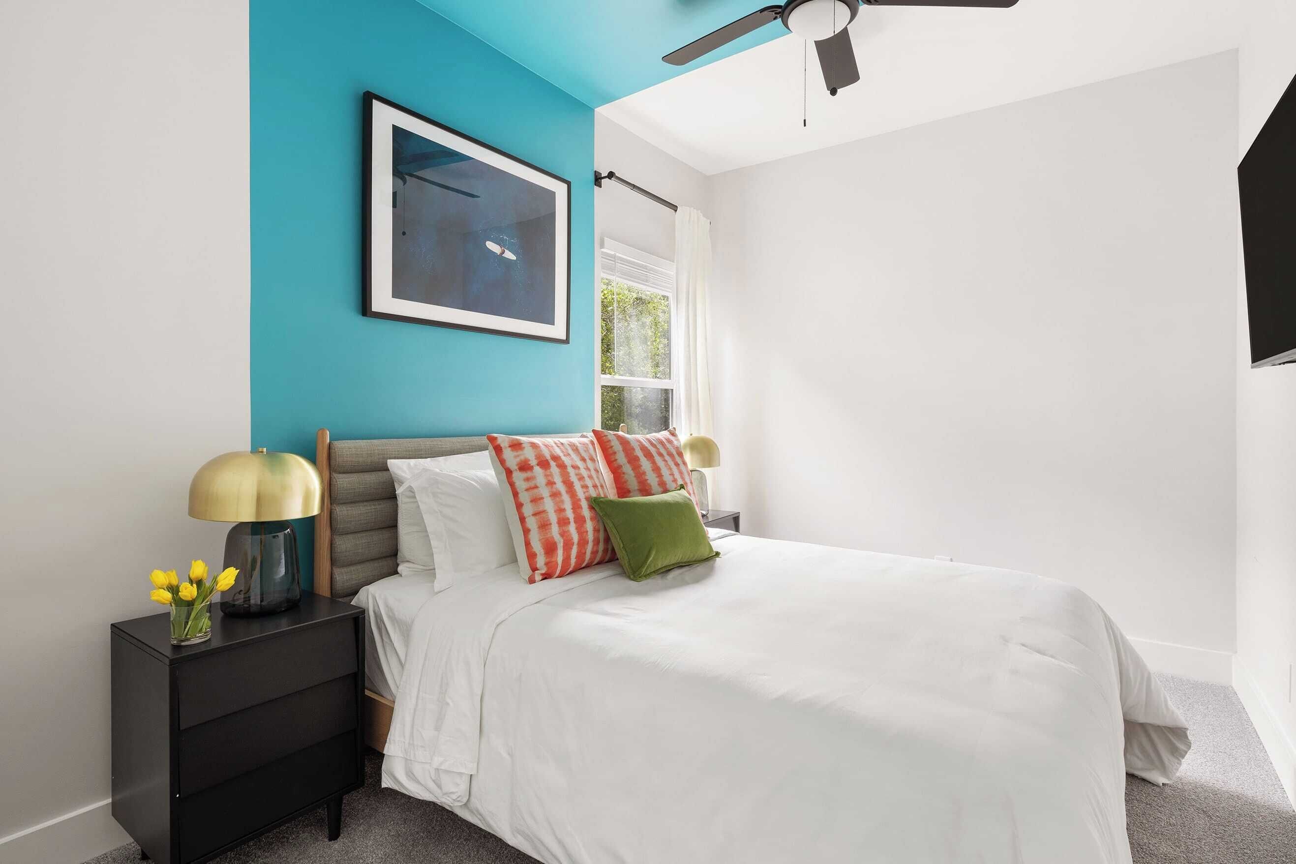 A bedroom with a bed, a nightstand, a ceiling fan, a window, and a framed picture on the wall.