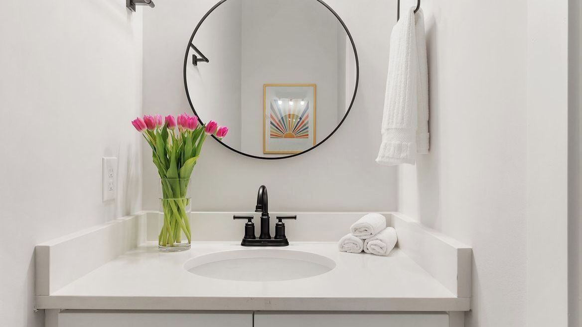 A bathroom with a white sink, a round mirror, a vase with pink flowers, and a towel rack with a white towel.