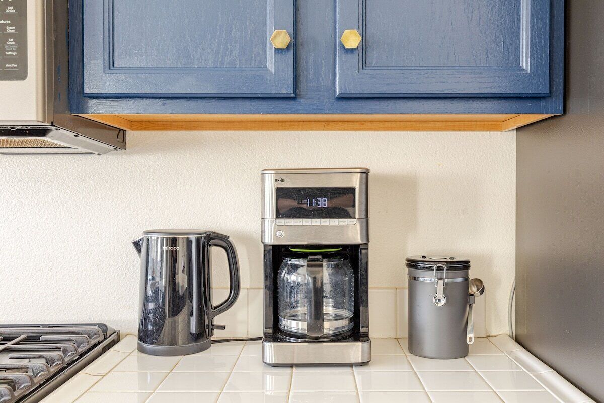 A kitchen with a coffee maker, a kettle, and a trash can.