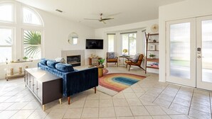 A spacious living room with a large sofa, a fireplace, and a television. There are large windows and a ceiling fan. - Spacious, Bright 4-Bedroom Home with Mountain Views, Private Pool, and Hot Tub  (Palm Springs)