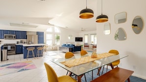 A modern kitchen with blue cabinets, a dining area with a glass table and yellow chairs, and a living space with a sofa and TV. - Spacious, Bright 4-Bedroom Home with Mountain Views, Private Pool, and Hot Tub  (Palm Springs)