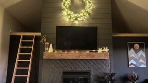 A living room with a fireplace, a TV mounted above it, a ladder, and a wreath on the wall.