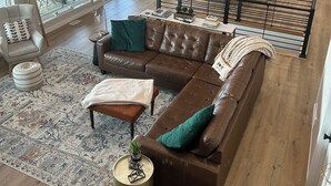 A spacious living area with a large sectional sofa, a coffee table, and a patterned rug. There is a dining area with a table and chairs, and a kitchen with modern appliances. The space has large windows and a staircase leading to an upper level.