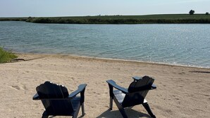 Two Adirondack chairs facing a body of water with a grassy bank and distant trees.