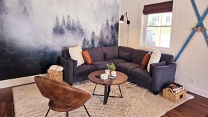 Un salón con un mural de un gran bosque, un sofá gris oscuro, un sillón de cuero marrón, una mesa de centro redonda y una ventana con persianas.