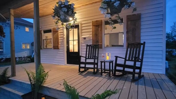 Una terraza de madera con dos sillas y una mesa, un arreglo floral colgante y una vela encendida en el porche de una casa blanca.