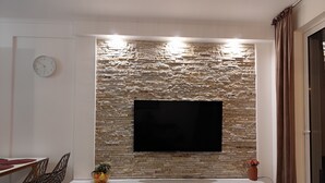 A modern living room with a stone wall, a flat-screen TV mounted on it, a white entertainment unit below, and a dining area with a table and chairs. - Golden Seaview Apartment  (Sarande)