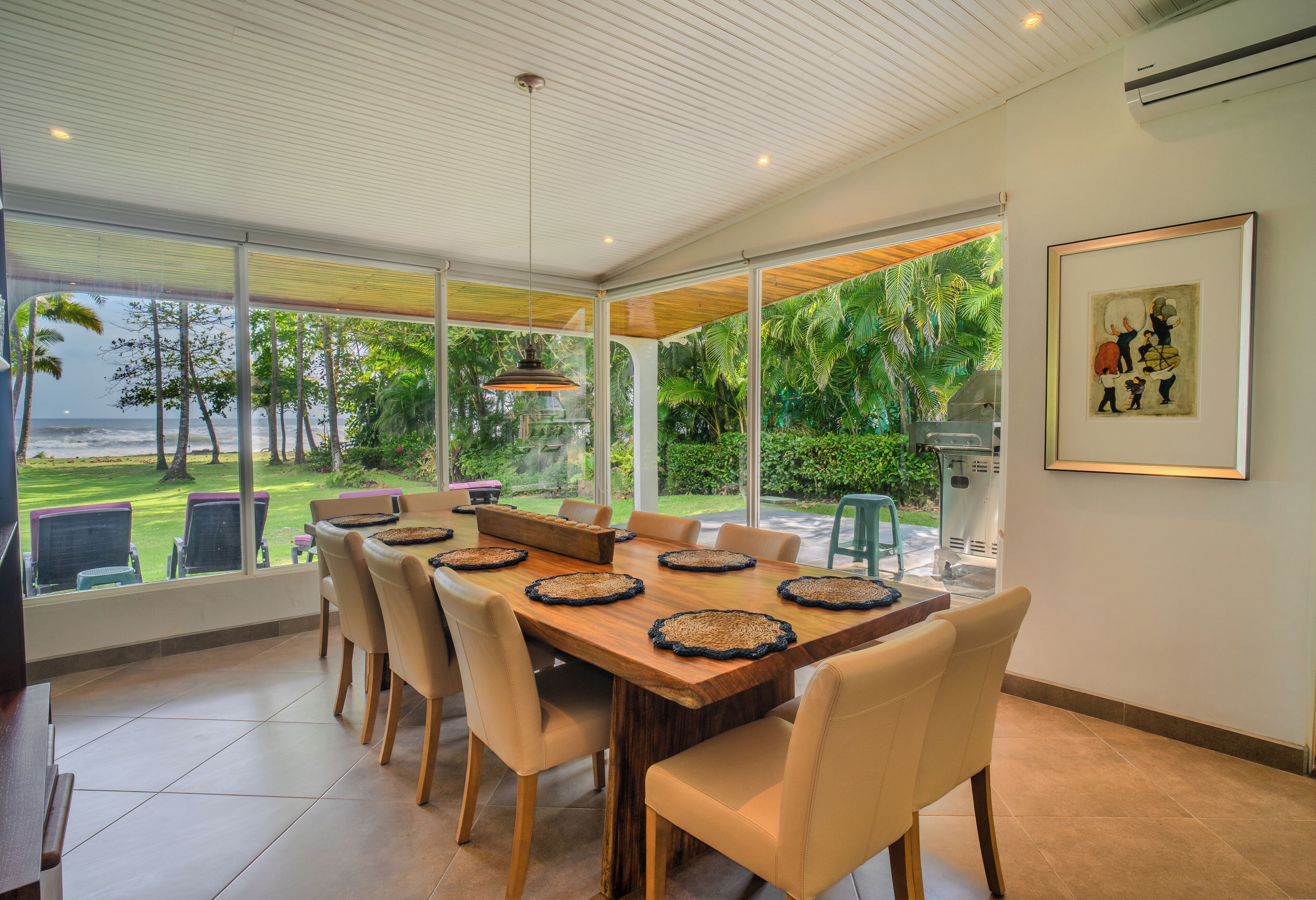 A spacious dining area with a large table and chairs, large windows offering a view of the outdoors, and a framed artwork on the wall.