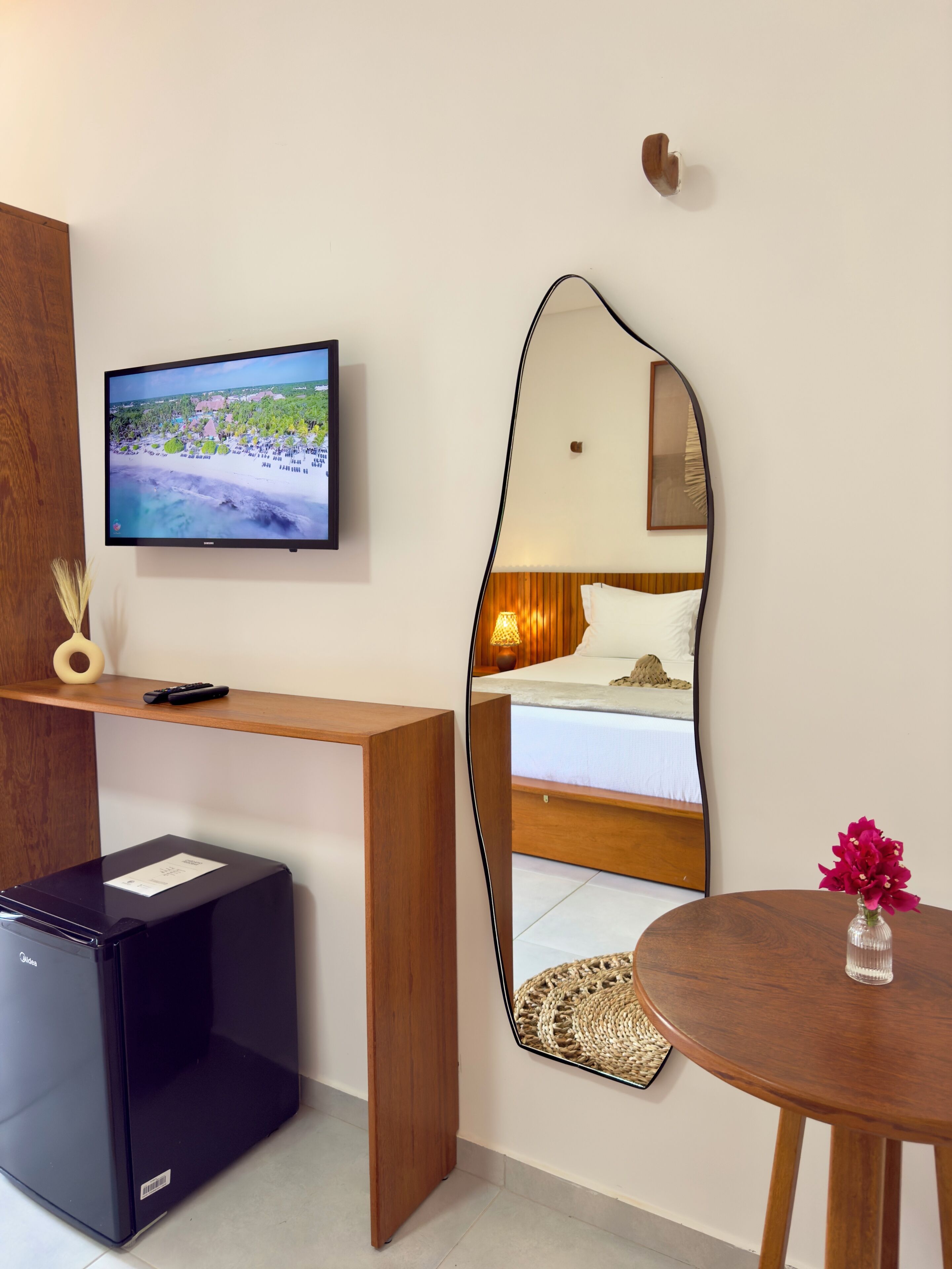 Une chambre d’hôtel équipée d’une télévision, d’un bureau en bois, d’un réfrigérateur noir, d’un grand miroir, d’un lit et d’une table ronde agrémentée d’un vase.