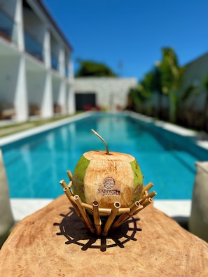 A poolside view with a wooden table holding a coconut with a straw, and a building in the background. - Yahweh Hotel  (Touros)