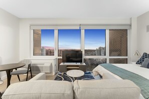 A modern hotel room with a large window, a flat-screen TV, a sofa, a small round table, and a bed with a blue blanket. - Cozy studio with fitness room, WiFi in lovely Queens (Queens)