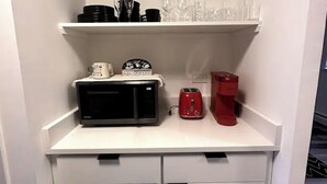 A kitchen with white cabinets, a microwave, a red coffee maker, and shelves with glassware and black mugs.