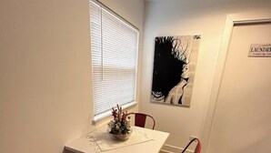 A dining area with a white table, red chairs, a window with blinds, a black abstract painting, and a sign reading "LAUNDRY".