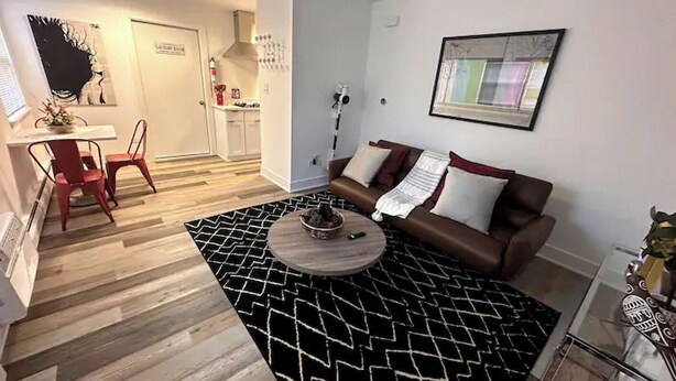 A modern living room with a brown sofa, a coffee table, and a black and white patterned rug. There is a dining area with a table and chairs, and a kitchen in the background.