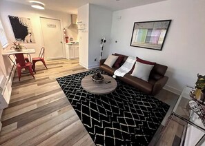Un salon moderne avec un canapé marron, une table basse et un tapis à motifs noir et blanc. On y trouve également un coin repas avec une table et des chaises, et une cuisine en arrière-plan.