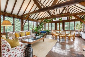 Una habitación amplia con techo alto, vigas de madera, un conjunto de sofás, una mesa de comedor con sillas y plantas en macetas.