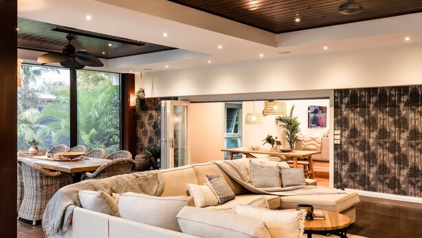 A spacious living area with a sofa, dining table, and chairs. There is a large window with a view of greenery outside. - Pet Friendly - Bellini Bliss with Gas Heated Pool (Tootgarook)