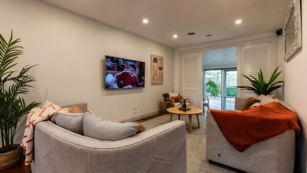 Ein modernes Wohnzimmer mit Fernseher, bequemen Sitzmöglichkeiten, einem kleinen Tisch und großen Fenstern.