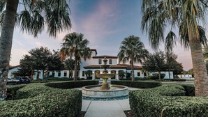 A well-manicured garden with a central fountain, palm trees, and a building with a tiled roof. - Entire Home in Kissimmee, FL - Windsor at Westside (Kissimmee)