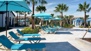 A poolside area with blue lounge chairs and umbrellas, surrounded by palm trees and buildings. - Entire Home in Kissimmee, FL - Windsor at Westside (Kissimmee)