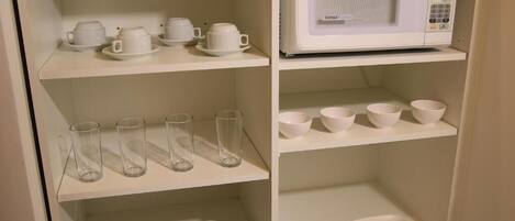 A kitchen with white shelves holding cups, glasses, bowls, and plates, and a white microwave on the countertop.