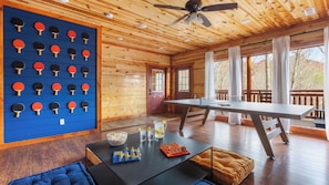 A wooden cabin interior with a ping pong table, a blue wall with ping pong paddles, a table with snacks, and a window with a view.