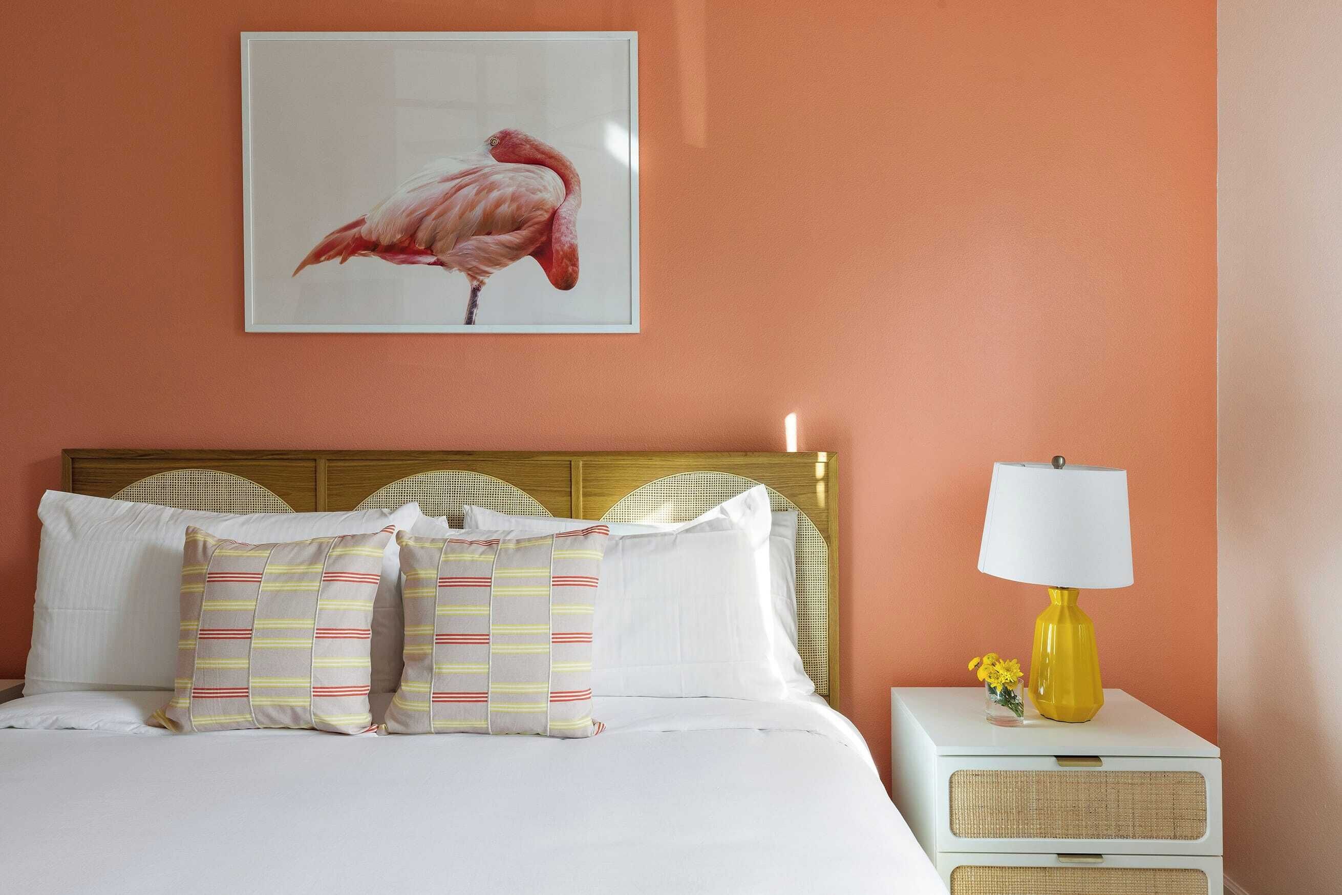 A bed with white bedding and striped pillows, a framed flamingo artwork on the wall, and a yellow lamp on a bedside table.