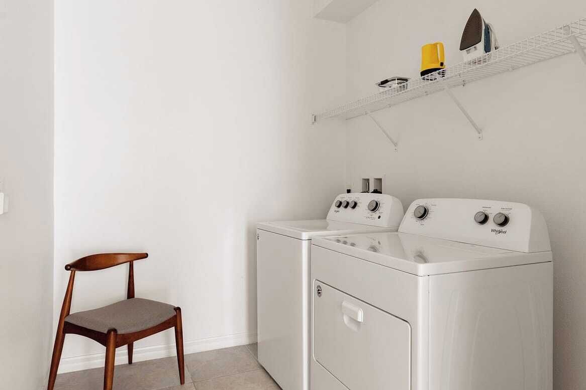 Une buanderie équipée de deux lave-linge et deux sèche-linge à chargement frontal, tous deux blancs, d’une chaise en bois et d’une étagère en métal placée au-dessus des sèche-linge, sur laquelle se trouvent un récipient jaune et un objet noir.