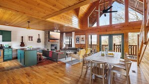 A spacious cabin interior with a kitchen, dining area, and living room, featuring wooden floors, a fireplace, and large windows. - Bears Den Way by Avantstay Private Golf Sim, Firepit & Mountain Views Near Dollywood (Sevierville)