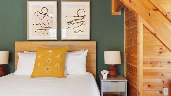Una cama bien hecha con sábanas blancas y una única almohada amarilla, flanqueada por dos mesitas de noche con lámparas y un pequeño jarrón con flores. Dos cuadros enmarcados que representan paisajes cuelgan sobre la cama.