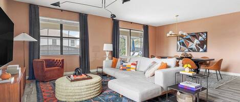 A modern living room with a sofa, ottoman, and coffee table, a dining area with chairs, and a TV on the left.