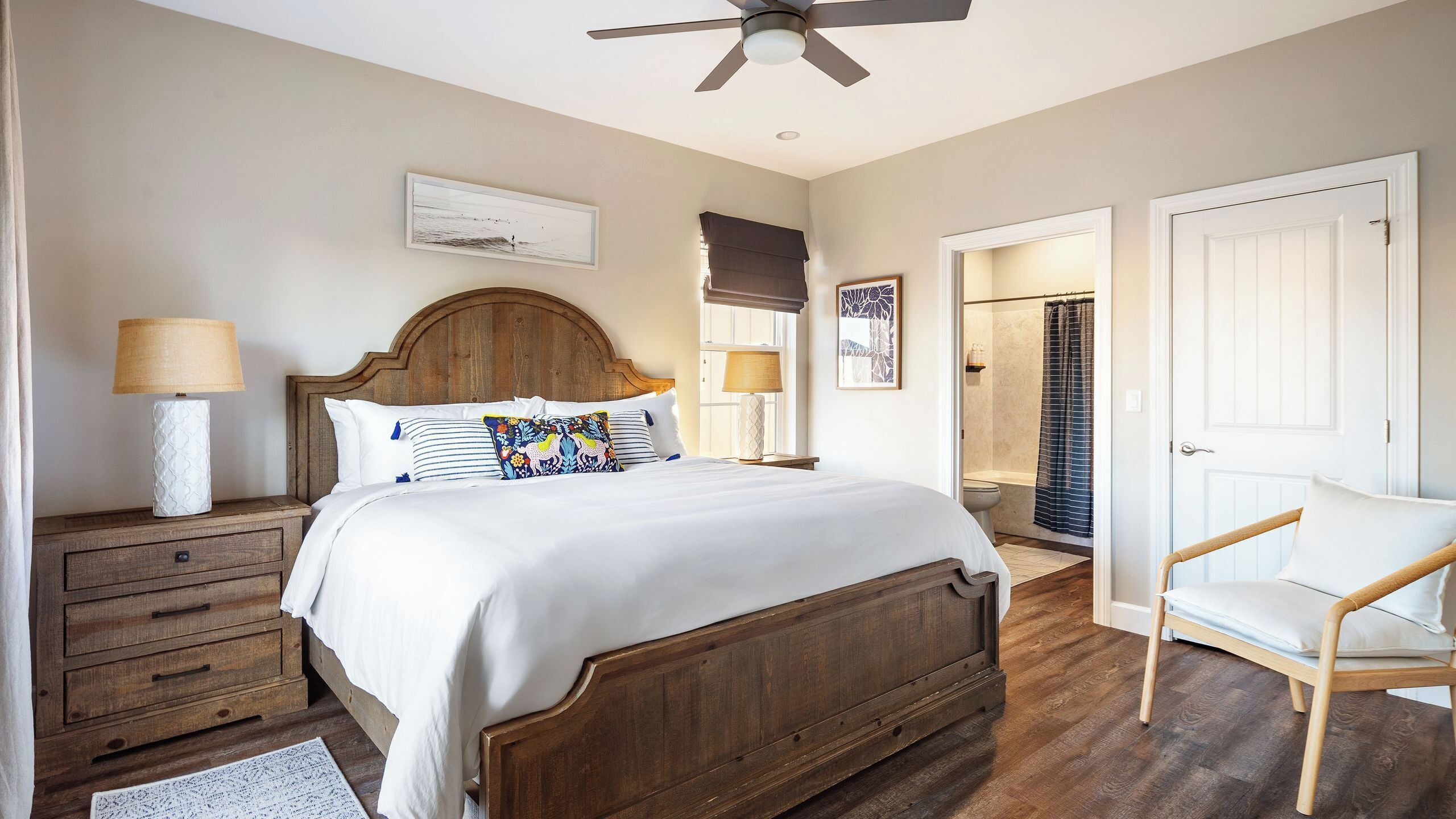 A bedroom with a wooden bed, two bedside tables with lamps, a chair, a ceiling fan, and a door leading to a bathroom.