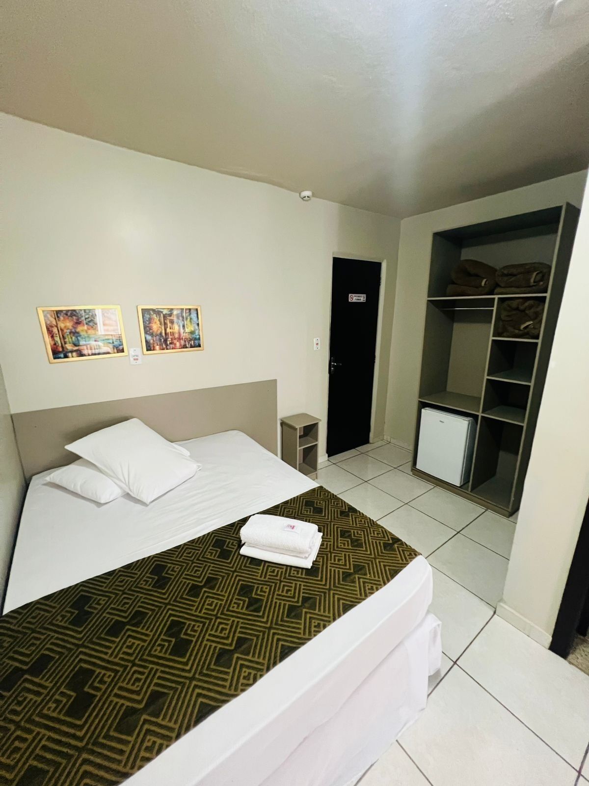 A hotel room with a bed, two framed pictures on the wall, a wardrobe, and a small cabinet.