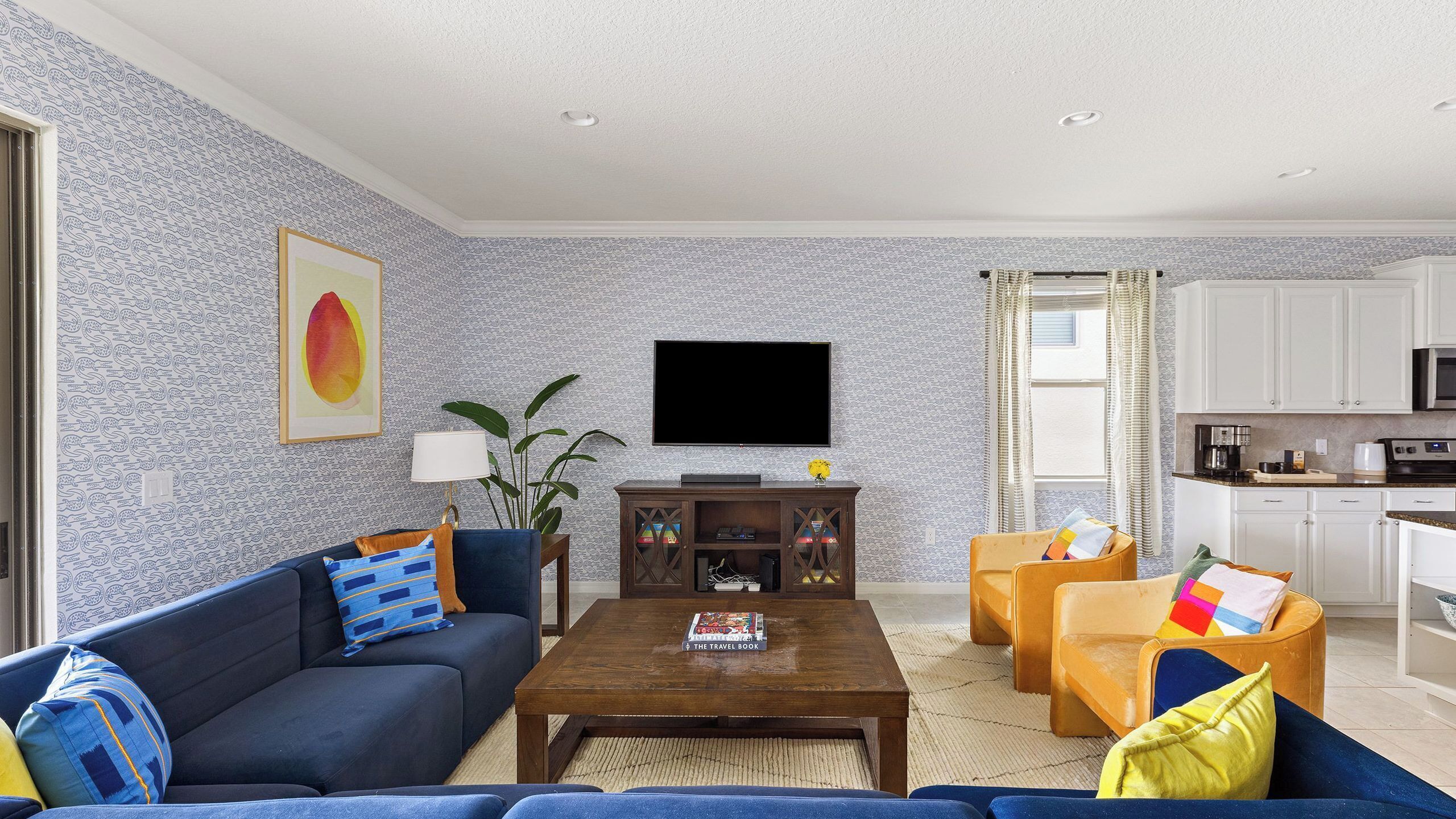 A living room with a sofa, coffee table, and TV. There is a kitchen area with cabinets and appliances.