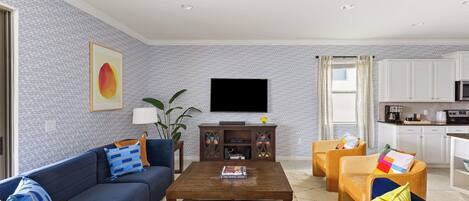 A living room with a sofa, coffee table, and TV. There is a kitchen area with cabinets and appliances.